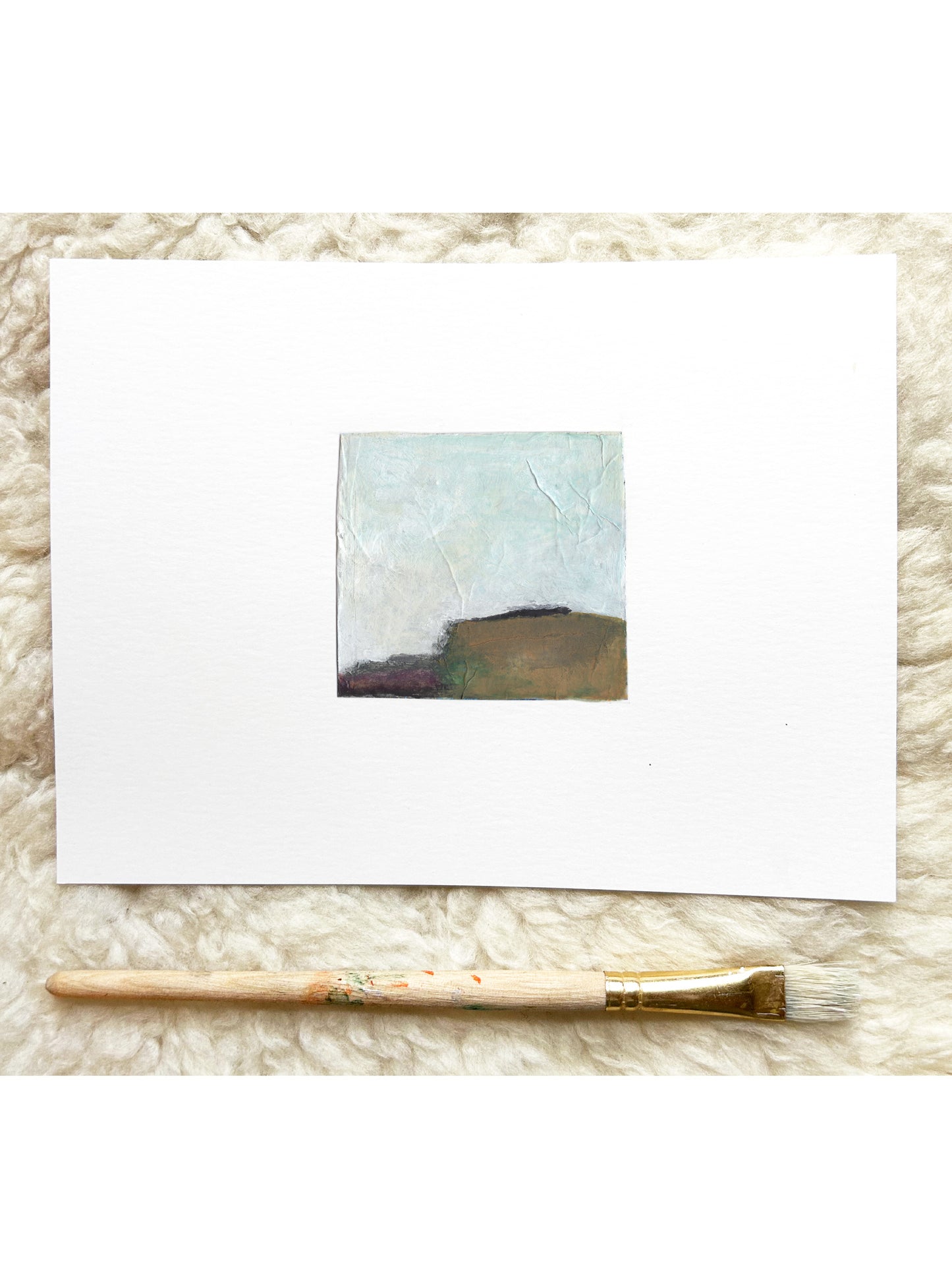 A small abstract landscape painting on a white card, displayed on a white textured rug with a paintbrush underneath.