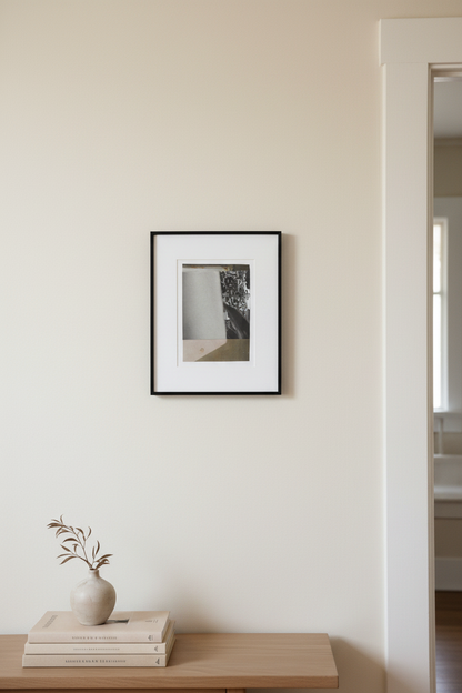 A minimalist black-framed art print with a white mat hangs on a warm beige wall above a light wood console table styled with a small ceramic vase and books.