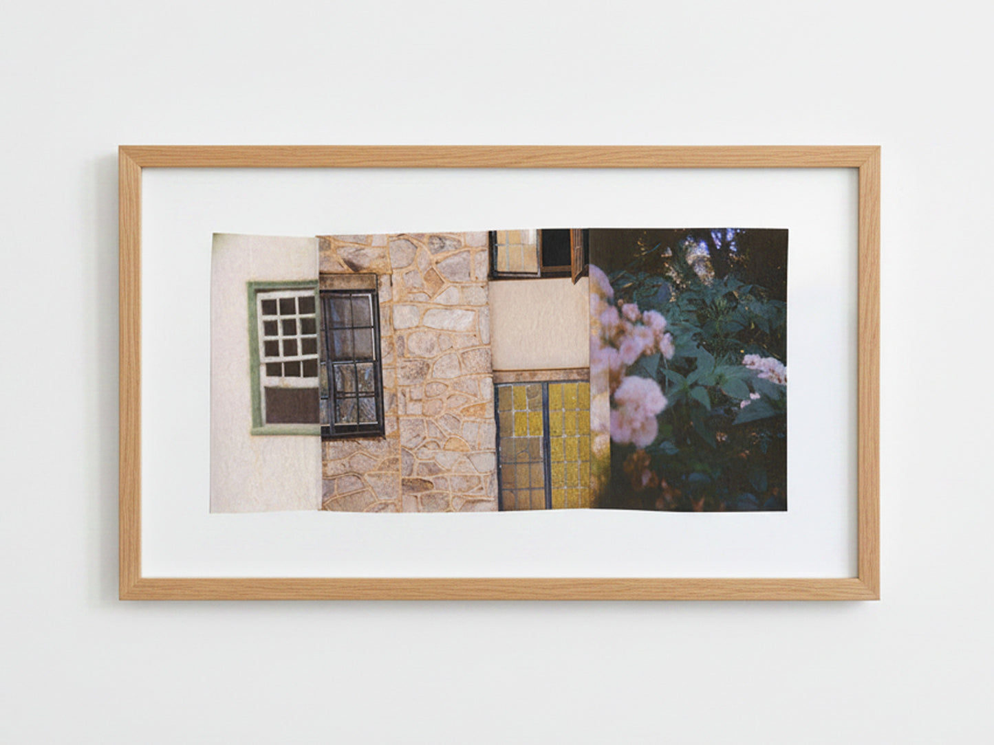 A framed art print featuring a street scene with a stone wall, windows, and pink flowers, displayed in a light wood frame against a white wall.