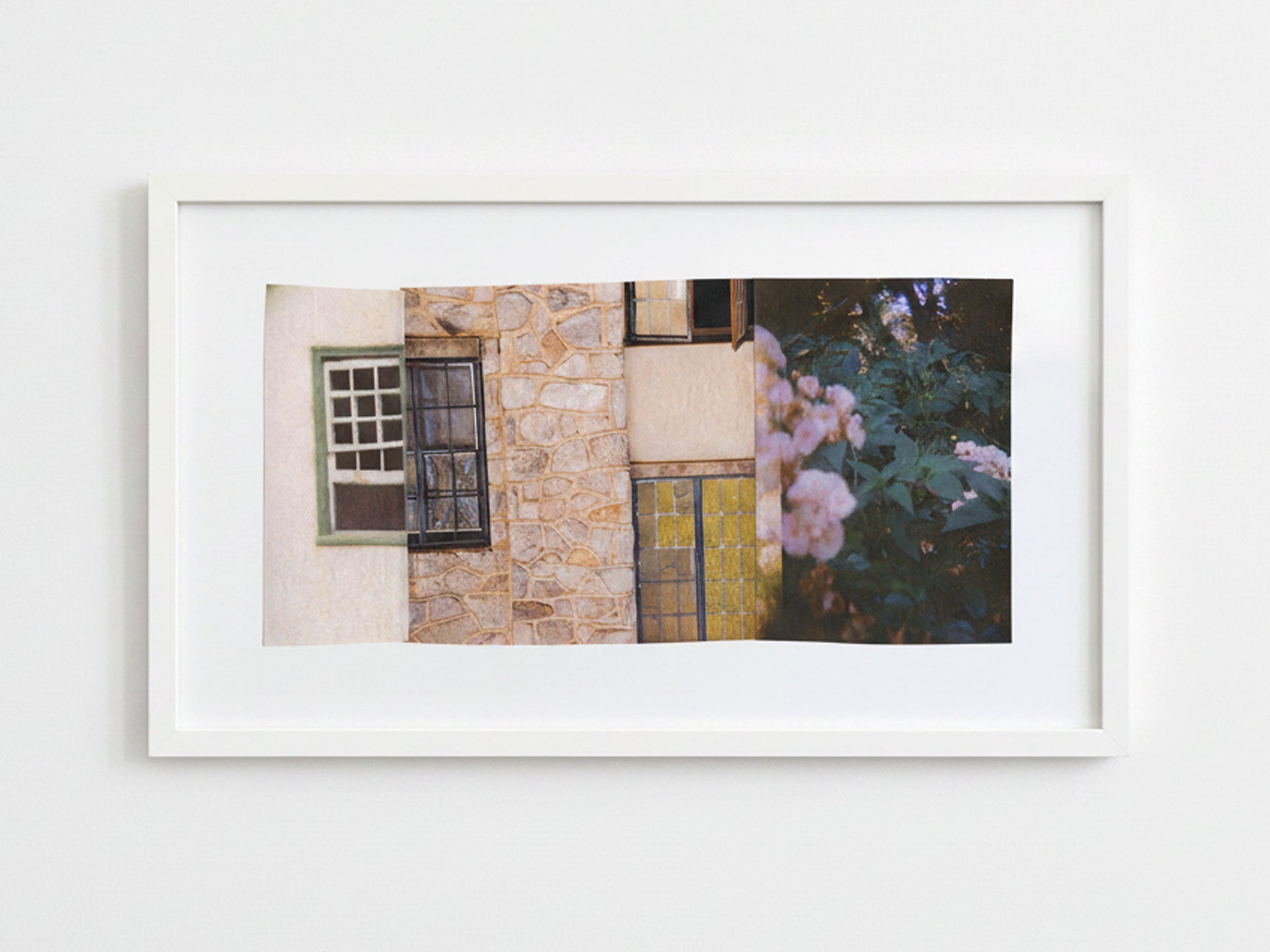 A framed horizontal art print featuring a rustic stone building facade with windows and pink flowers, displayed on a clean white wall.
