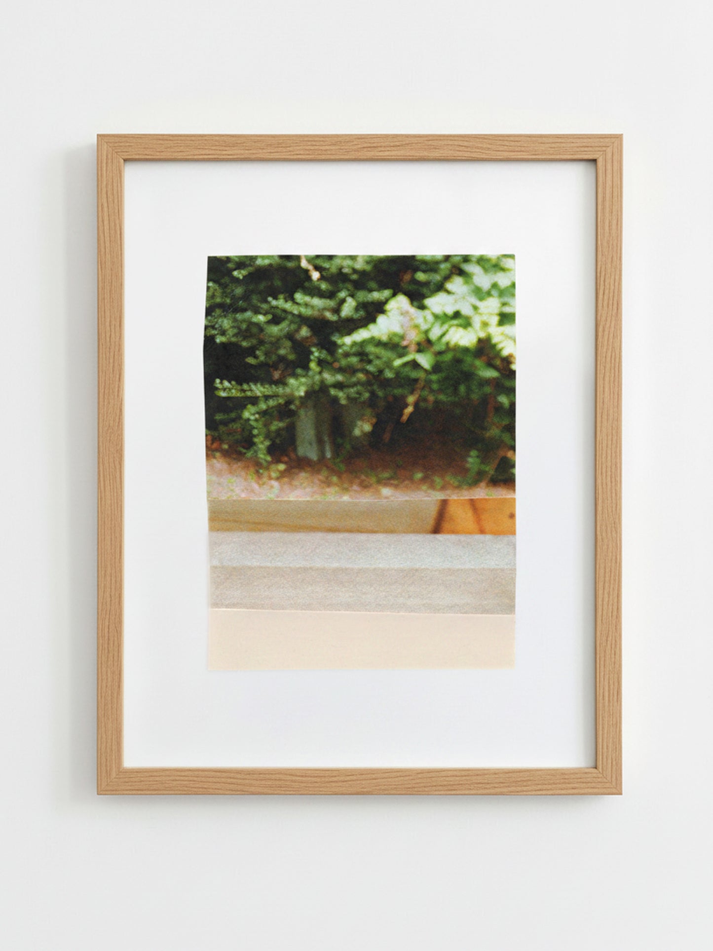 A framed art print featuring a photograph of green foliage and a wooden planter, mounted with a white mat in a light oak wood frame against a white wall.