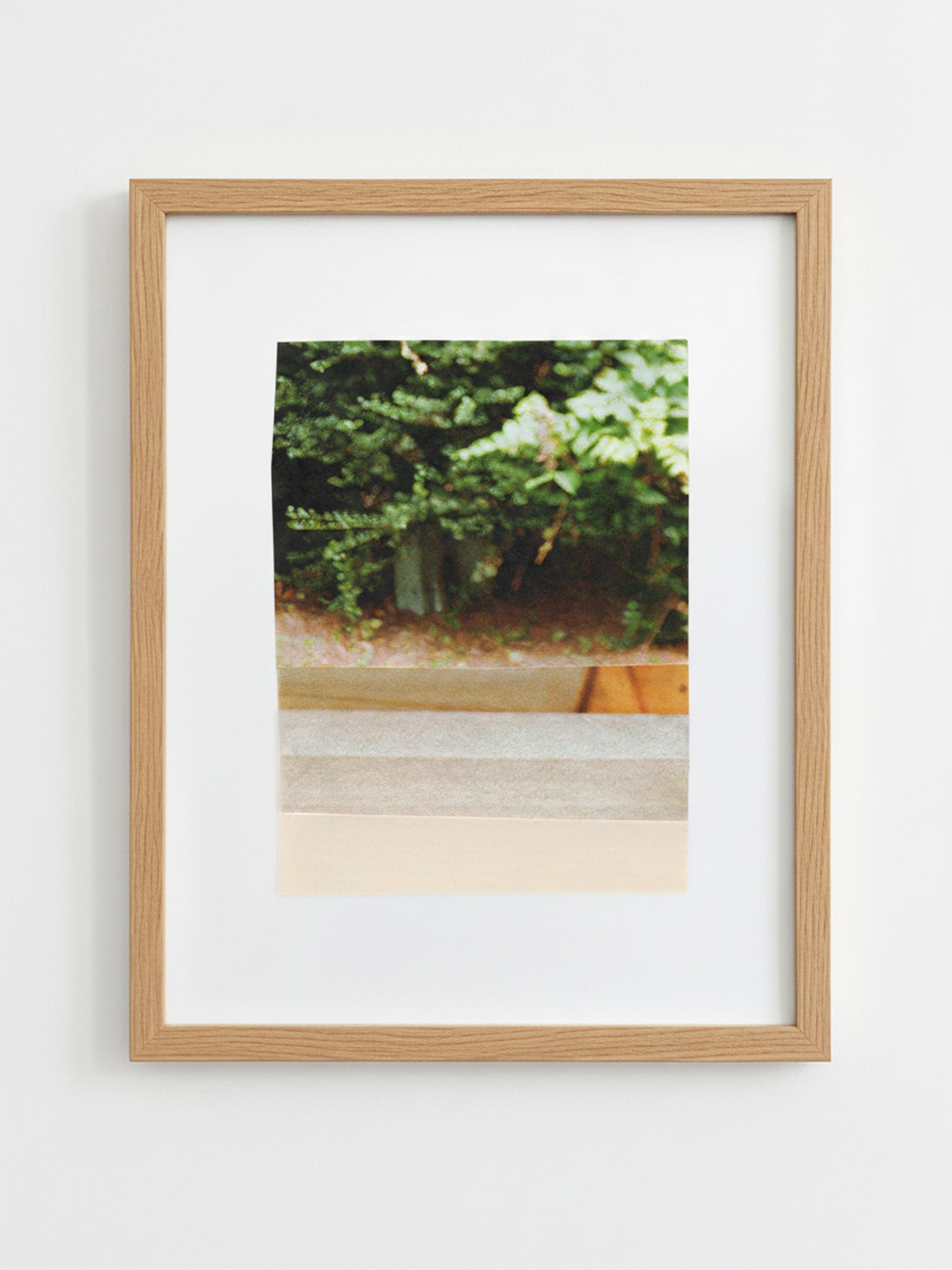 A framed art print featuring a photograph of green foliage and a wooden planter, mounted with a white mat in a light oak wood frame against a white wall.