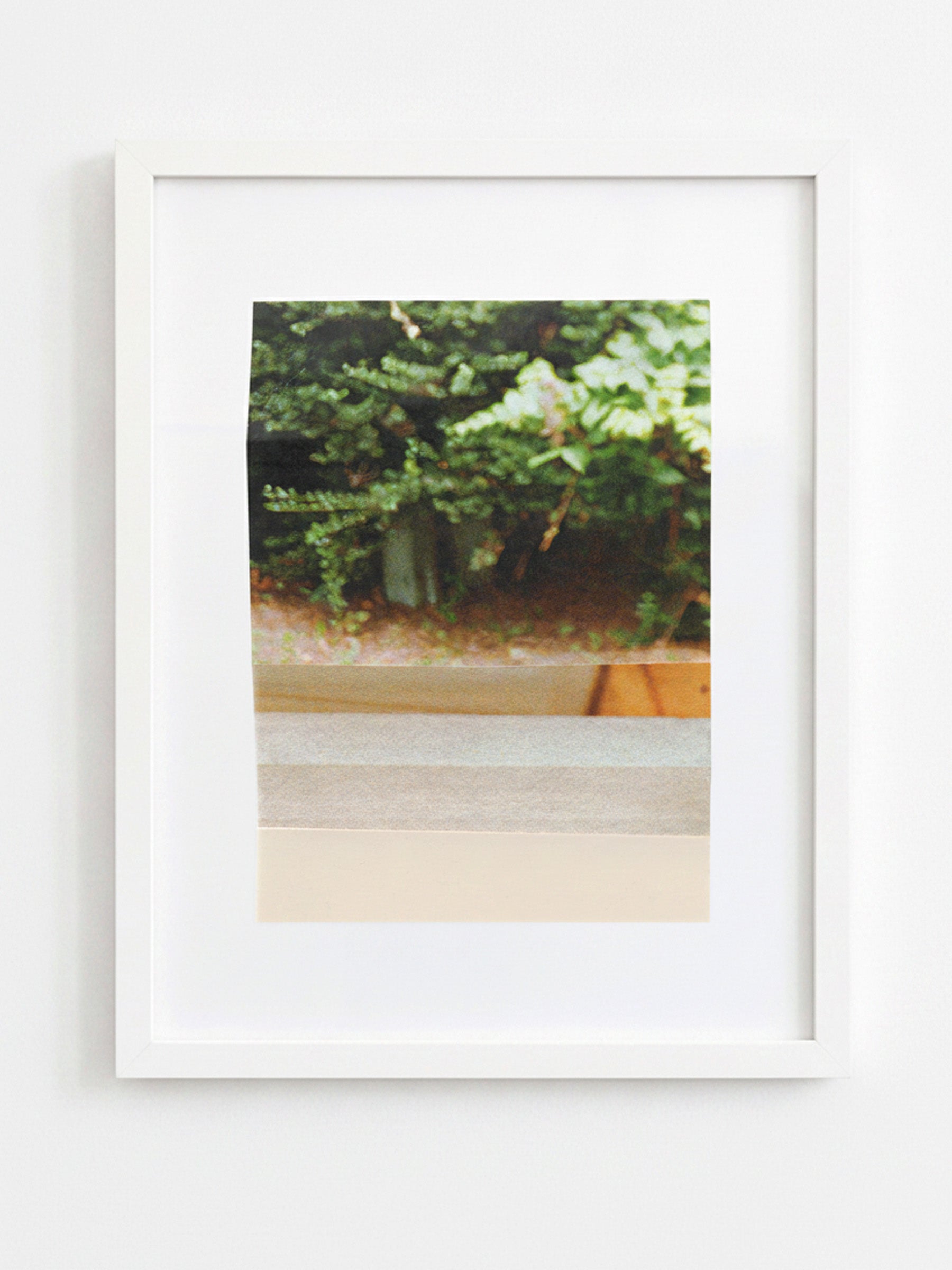 A framed art print featuring a soft-focus photograph of green foliage and a beige wall, presented in a white frame against a white wall.