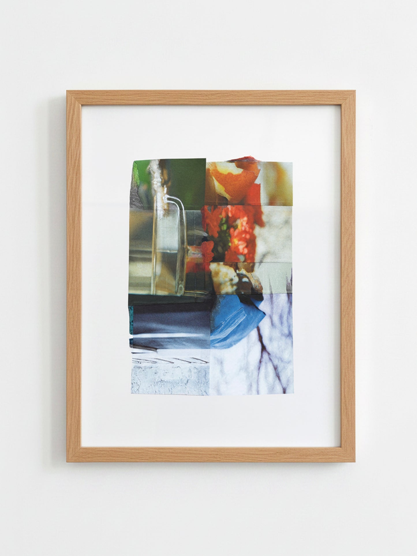 A framed abstract collage art print featuring a mix of photographic and painterly elements in green, orange, blue, and white, set in a light natural wood frame with a white mat, hanging on a plain white wall.