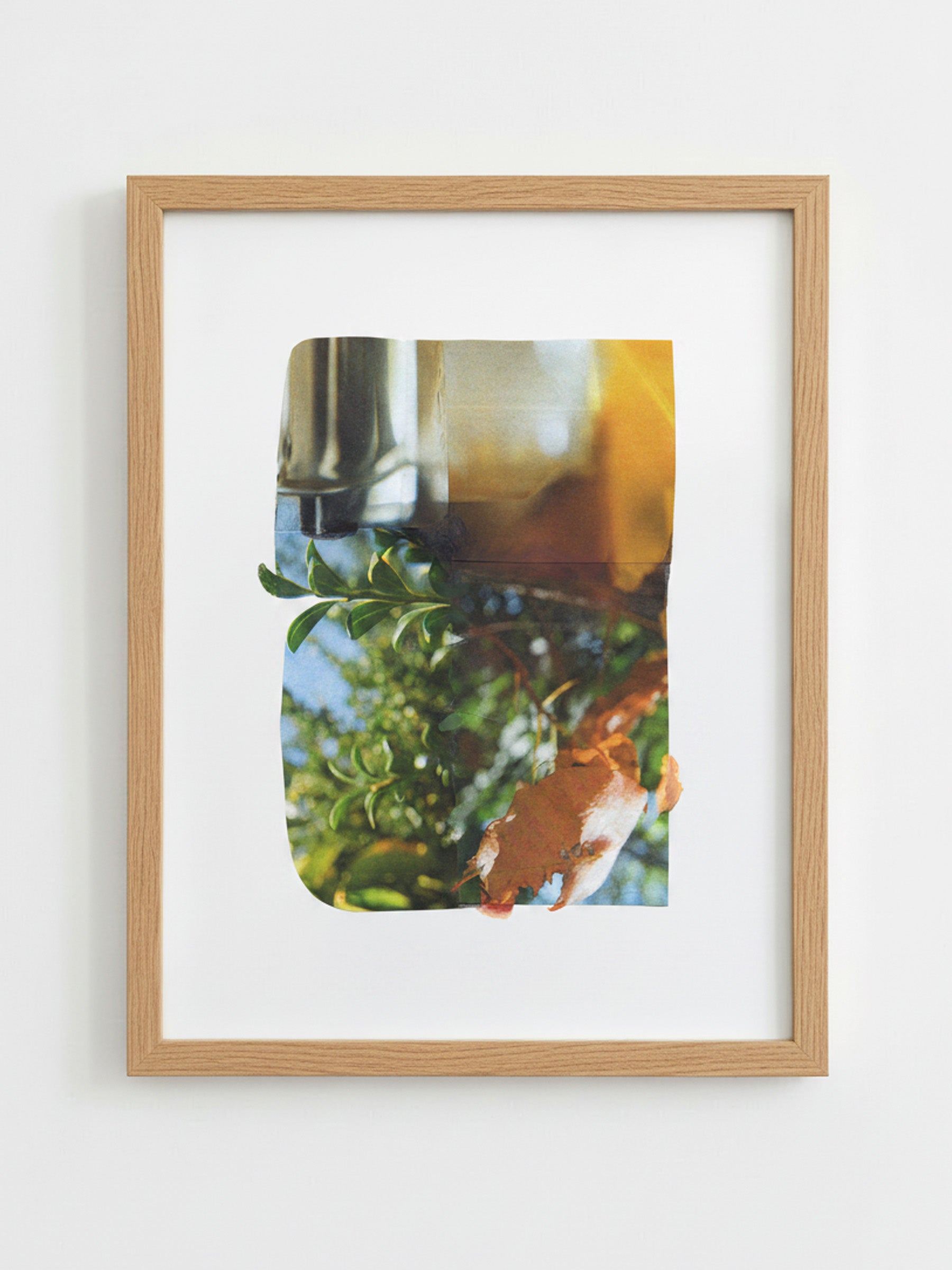 A framed art print featuring an abstract collage of green foliage, a brown textured shape, and warm yellow/orange tones, set in a light natural wood frame with a white mat against a white wall.