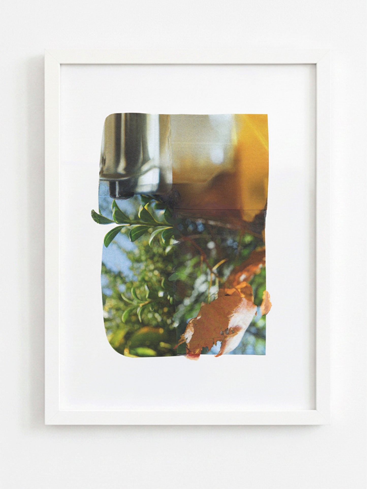 A framed art print featuring an abstract photographic composition with green foliage, a metallic silver object, and warm amber and orange tones, displayed in a white frame against a light gray wall.
