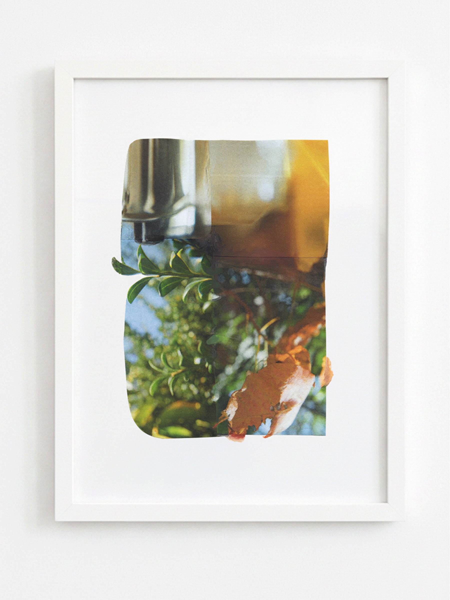 A framed art print featuring an abstract photographic composition with green foliage, a metallic silver object, and warm amber and orange tones, displayed in a white frame against a light gray wall.