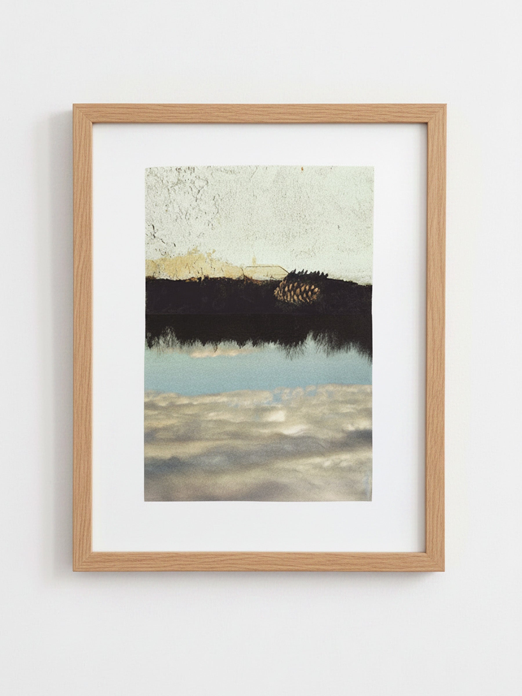 A framed art print featuring an abstract landscape with a dark horizontal band, a pale sky, and reflective water, presented in a light natural wood frame with a white mat against a white wall.