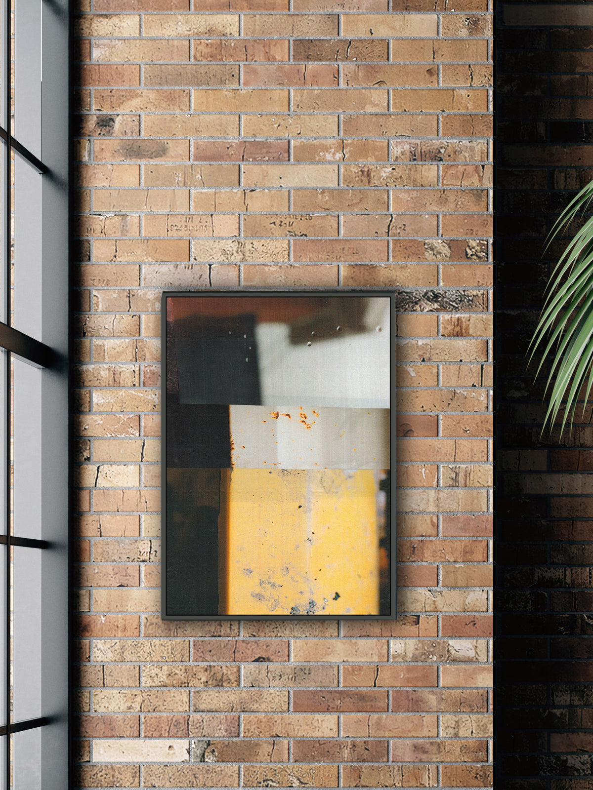 A framed abstract art print featuring geometric blocks of yellow, white, and dark gray, mounted on a rustic brick wall next to a window and a green plant.