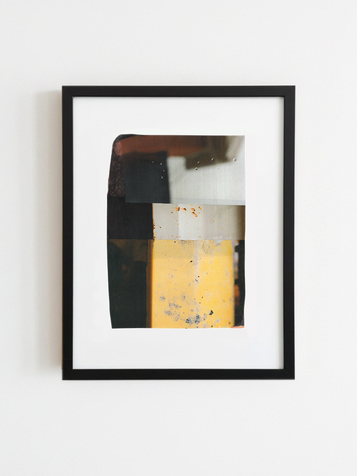 A framed abstract art print featuring geometric blocks of yellow, black, white, and brown, presented in a black frame against a plain white wall.