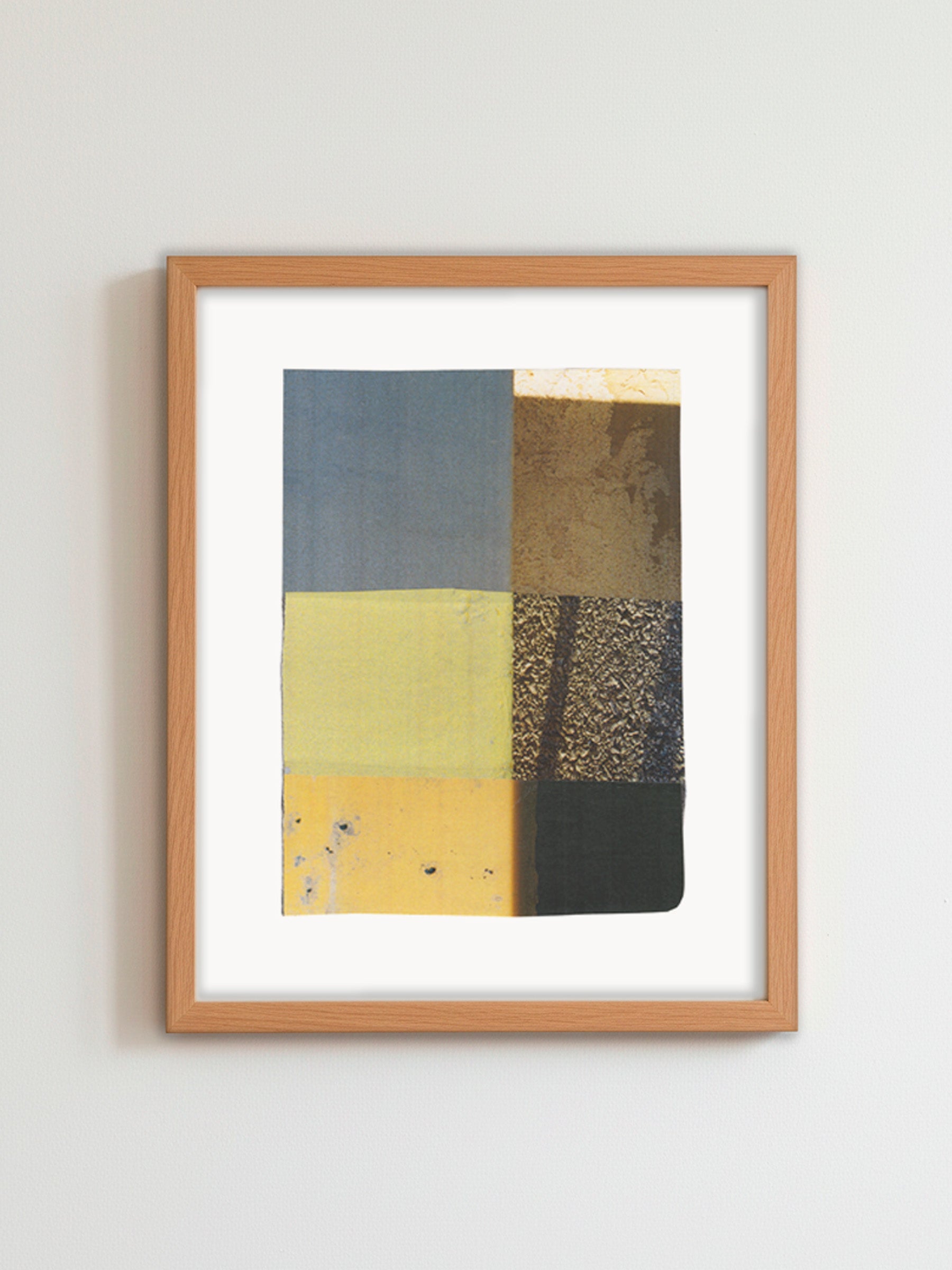 A framed abstract art print featuring a grid of textured blocks in slate blue, mustard yellow, black, and metallic gold, housed in a light oak frame and mounted on a neutral light gray wall.