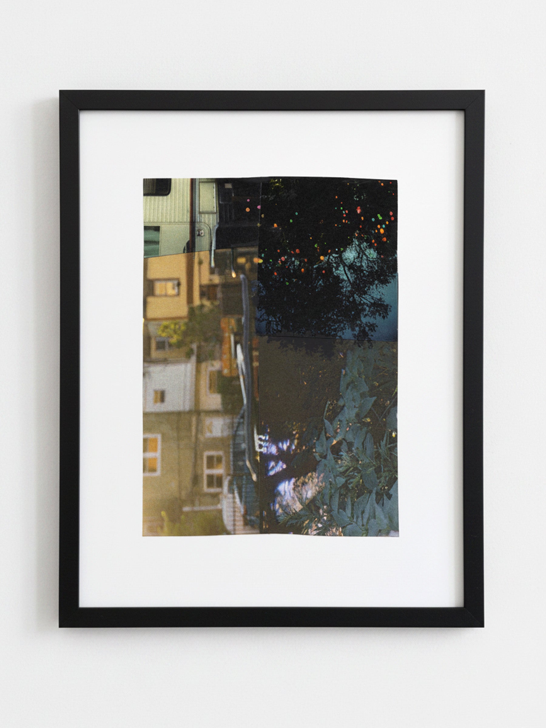 A framed art print featuring an upside-down photograph of a street scene with buildings, trees, and reflections, presented in a black frame with a white mat against a light gray wall.