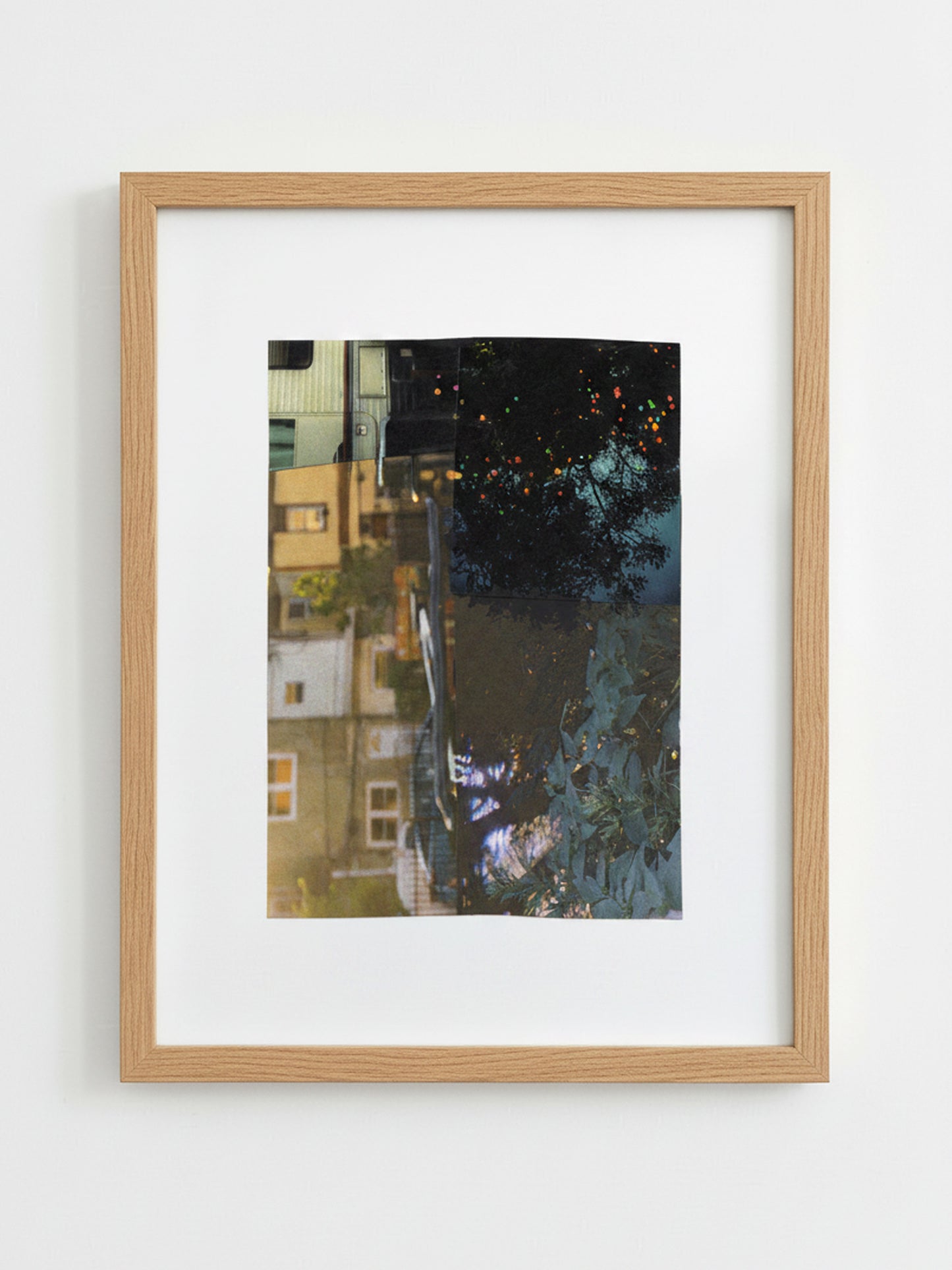 A framed art print featuring a vertical photograph of a city street reflection, set in a light oak wood frame, hanging on a plain white wall.