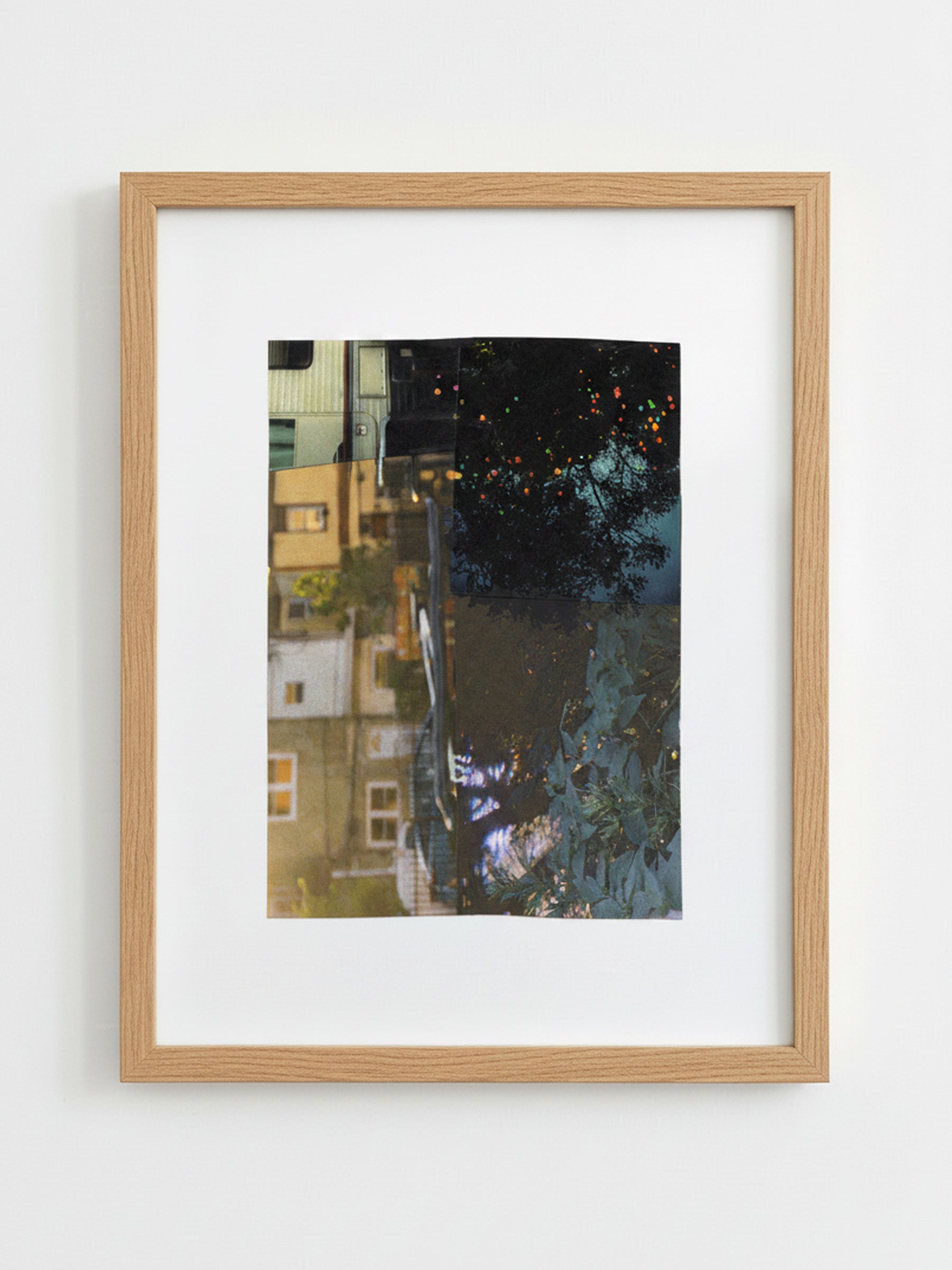 A framed art print featuring a vertical photograph of a city street reflection, set in a light oak wood frame, hanging on a plain white wall.