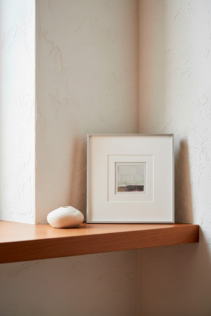 A small square framed artwork with a white mat and silver frame sits on a wooden shelf next to a smooth white stone vessel, set against a textured cream wall.