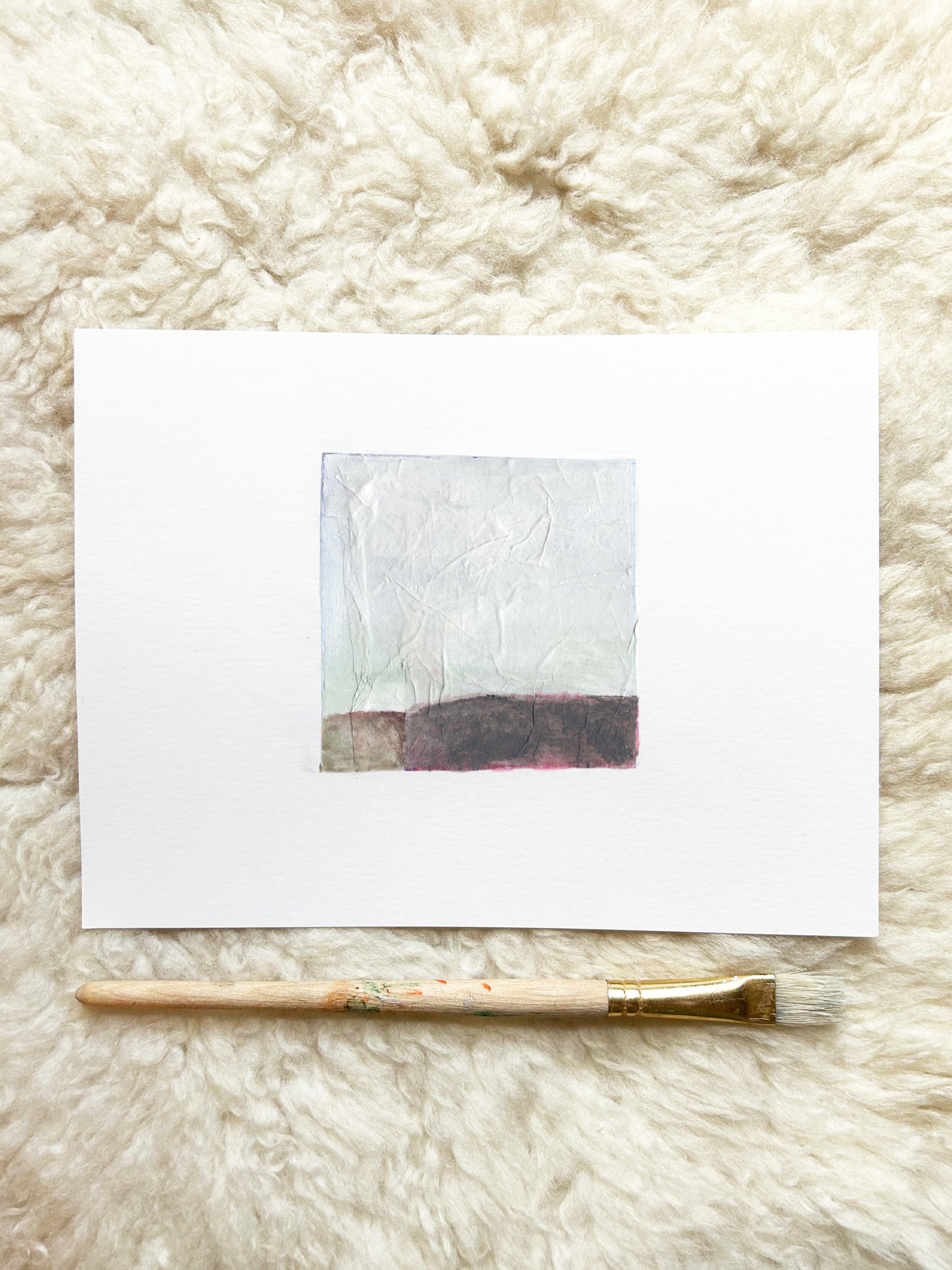 A small abstract art print on white paper, featuring a textured light gray upper section and a dark charcoal-brown band at the bottom, displayed on a fluffy cream rug with a paintbrush underneath.