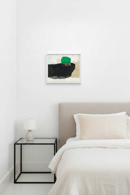 A framed abstract art print featuring a black block, beige background, and a green geometric shape, displayed on a white wall above a bed with a beige headboard and white bedding in a modern bedroom setting.